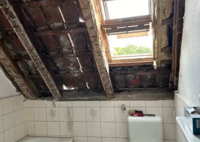 Badezimmer mit beschädigtem Dach und sichtbarem Fenster, Fliesenboden und Sanitäranlagen.